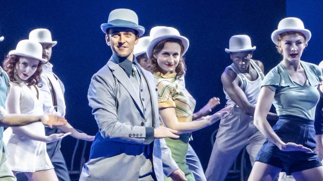 George Blagden (Bob) and members of the Company in Irving Berlin’s White Christmas. Photo by Johan Persson. Dancers lit in pale blue all wear bowler hats and perform in unison. They stretch one leg out and lean sideways with the other.