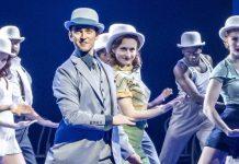 George Blagden (Bob) and members of the Company in Irving Berlin’s White Christmas. Photo by Johan Persson. Dancers lit in pale blue all wear bowler hats and perform in unison. They stretch one leg out and lean sideways with the other.